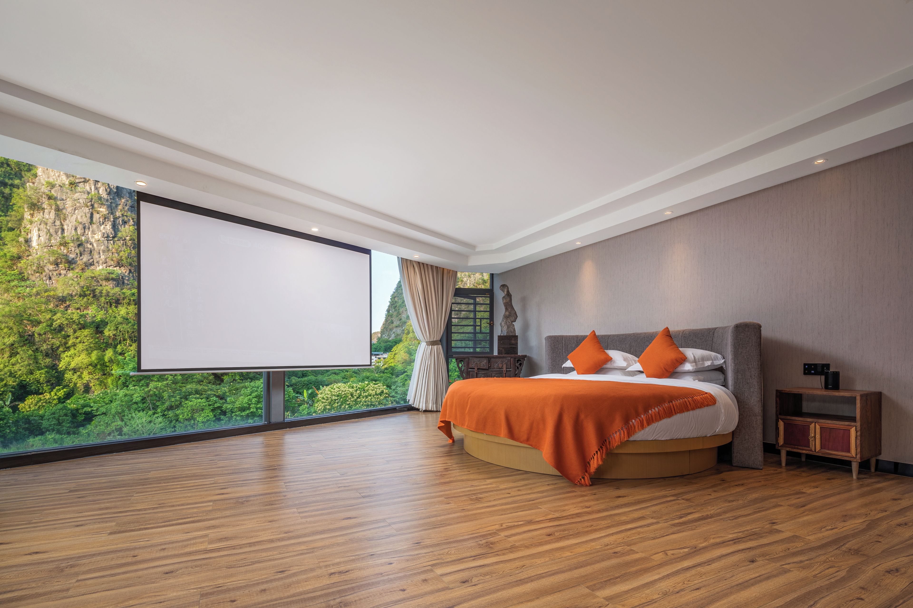 A modern bedroom with a large bed, a wooden floor, a projector screen, and a view of greenery outside.