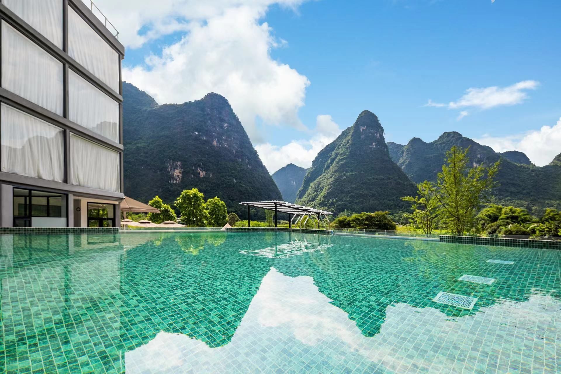 Una piscina con montañas al fondo, un edificio con amplias ventanas y cielo despejado.