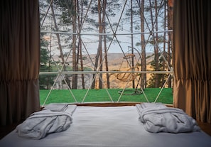 Una cama con ropa de cama y toallas blancas, una ventana con vista a un bosque y un conjunto de columpios.