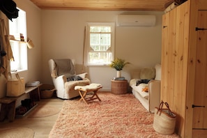Ein gemütliches Zimmer mit Holzbalkendecke, einem großen Fenster, einem weißen Sessel, einem kleinen Holzschemel, einem Weidenbeistelltisch und einem flauschigen Teppich.