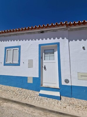 A building with a white and blue facade, a tiled roof, a white door with a window, and a small step leading to the entrance. - Casa de Santa Margarida (Mourão)