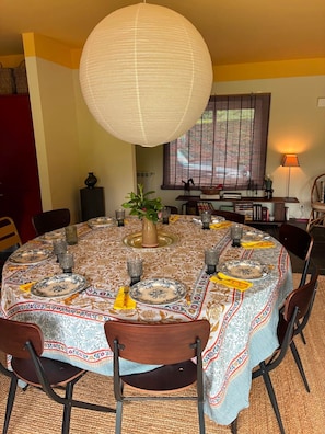 A dining area with a round table set for four, surrounded by chairs, a hanging light fixture, and a window with blinds. - Casa de Santa Eulália (Candemil e Gondar)