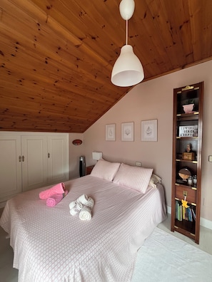 Quarto com teto de madeira, cama com roupa de cama rosa, luminária branca e estante com diversos objetos.