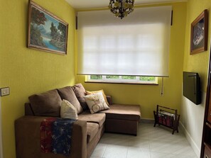 Ein Wohnzimmer mit einem braunen Sofa, einem Fenster mit Jalousien, einer Chandelier und einem Bild an der Wand.
