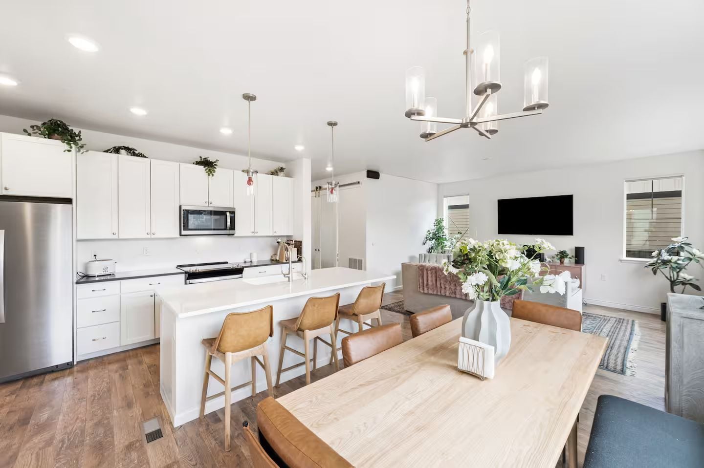 Een moderne keuken met witte kasten, een centraal kookeiland met barstoelen, een eettafel en een grote kroonluchter.