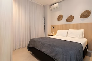 A hotel room with a bed, a nightstand, a wall-mounted air conditioner, and a window with white curtains. - South Beach - Copacabana (Rio de Janeiro)