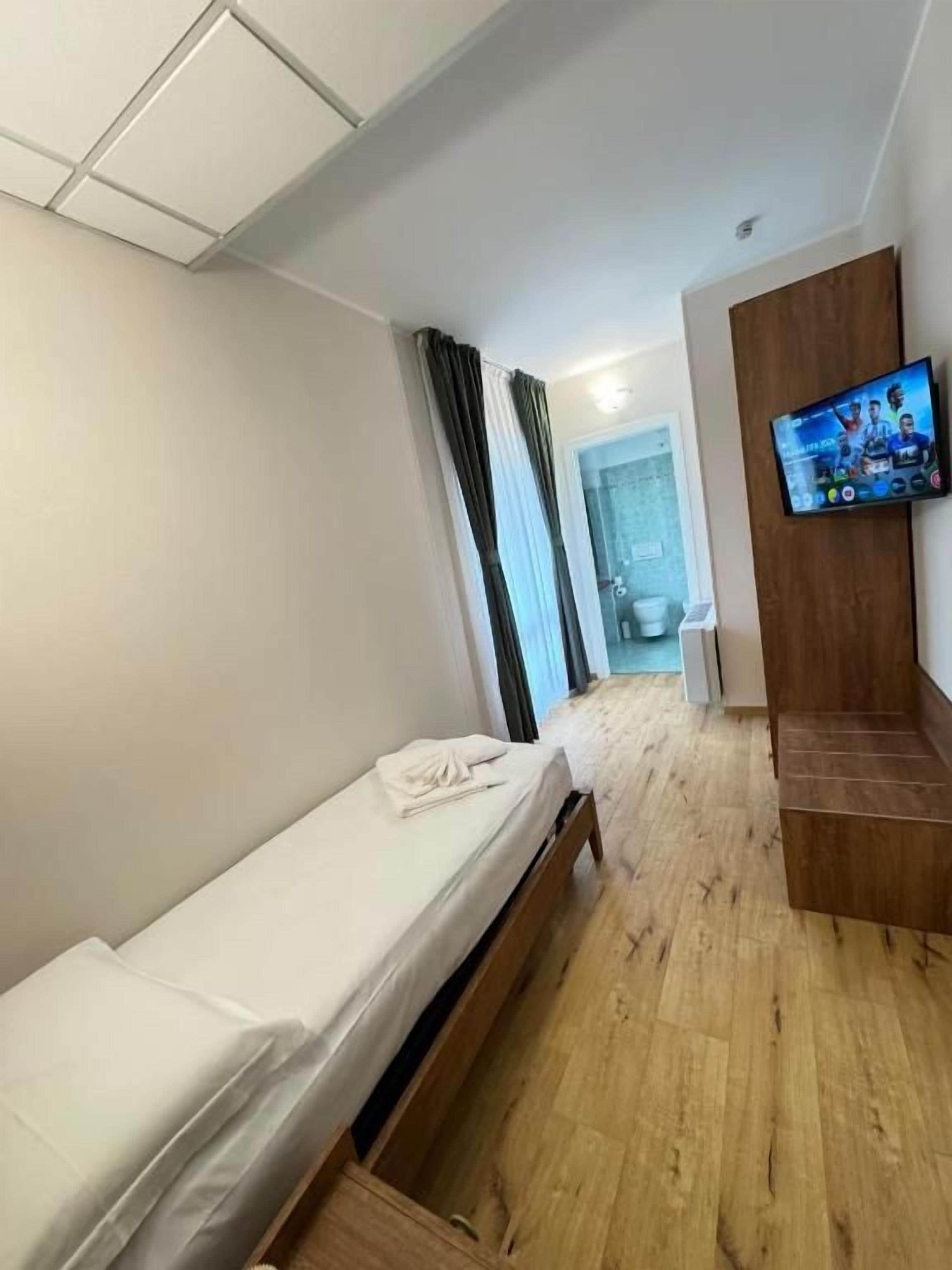 Een hotelkamer met een eenpersoonsbed, een houten vloer, een aan de muur bevestigde televisie en een raam met gordijnen.