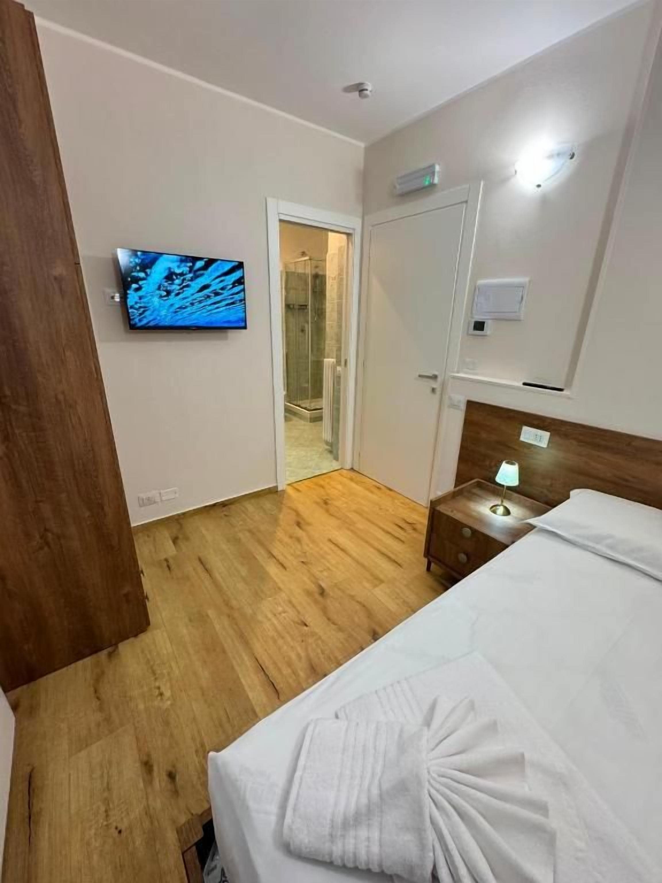 Ein Hotelzimmer mit Holzboden, einem Bett mit weißer Bettwäsche, einem an der Wand befestigten Fernseher und einer Tür zu einem Badezimmer.