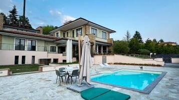 Ein Swimmingpool mit einer Steinterrasse, einem überdachten Sitzbereich und einem zweigeschossigen Gebäude im Hintergrund.