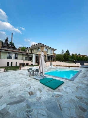 A swimming pool with a stone patio, a covered seating area, and a two-story building in the background.