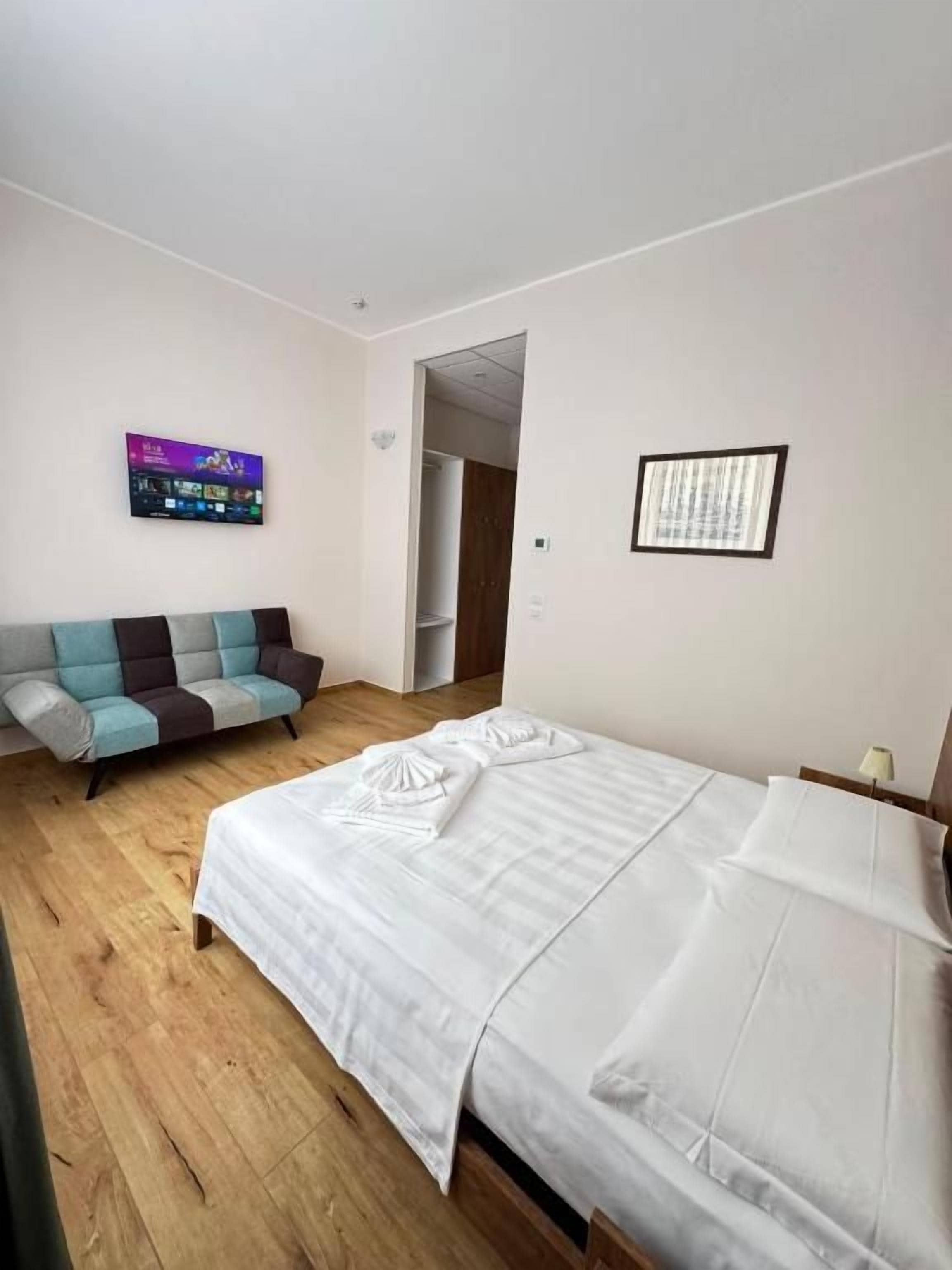 A hotel room with a bed, a sofa, a wall-mounted TV, and a door leading to another room.