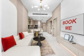 A modern living room with a sofa, a coffee table, and a TV screen displaying 'BOOK ME'. - Stayshort - Elegant 1 Bedroom in JVC That Sleeps 2 (Dubai)