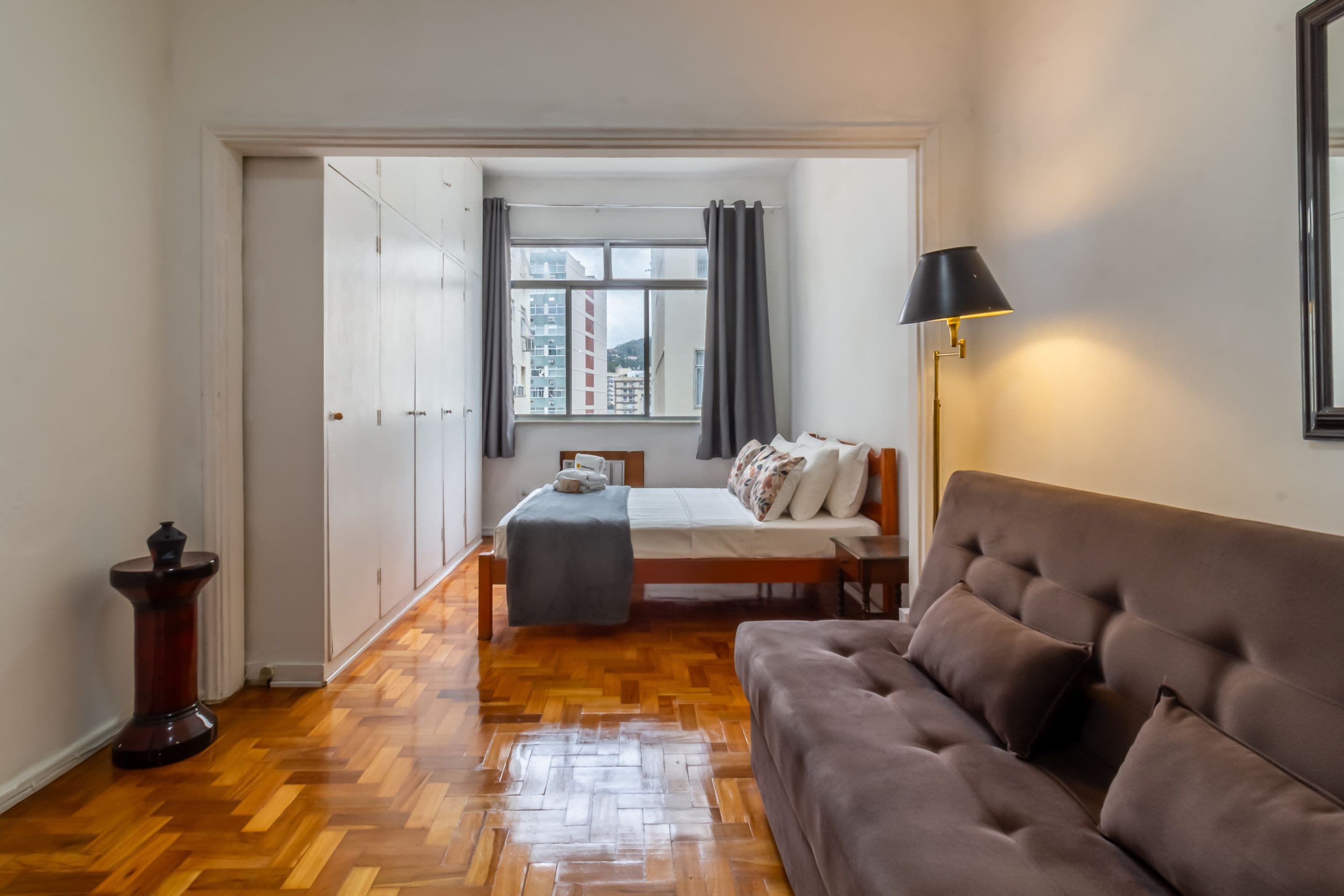 Une chambre à coucher comprenant un lit, une table de chevet, une lampe, une armoire et une vue sur des immeubles par la fenêtre.