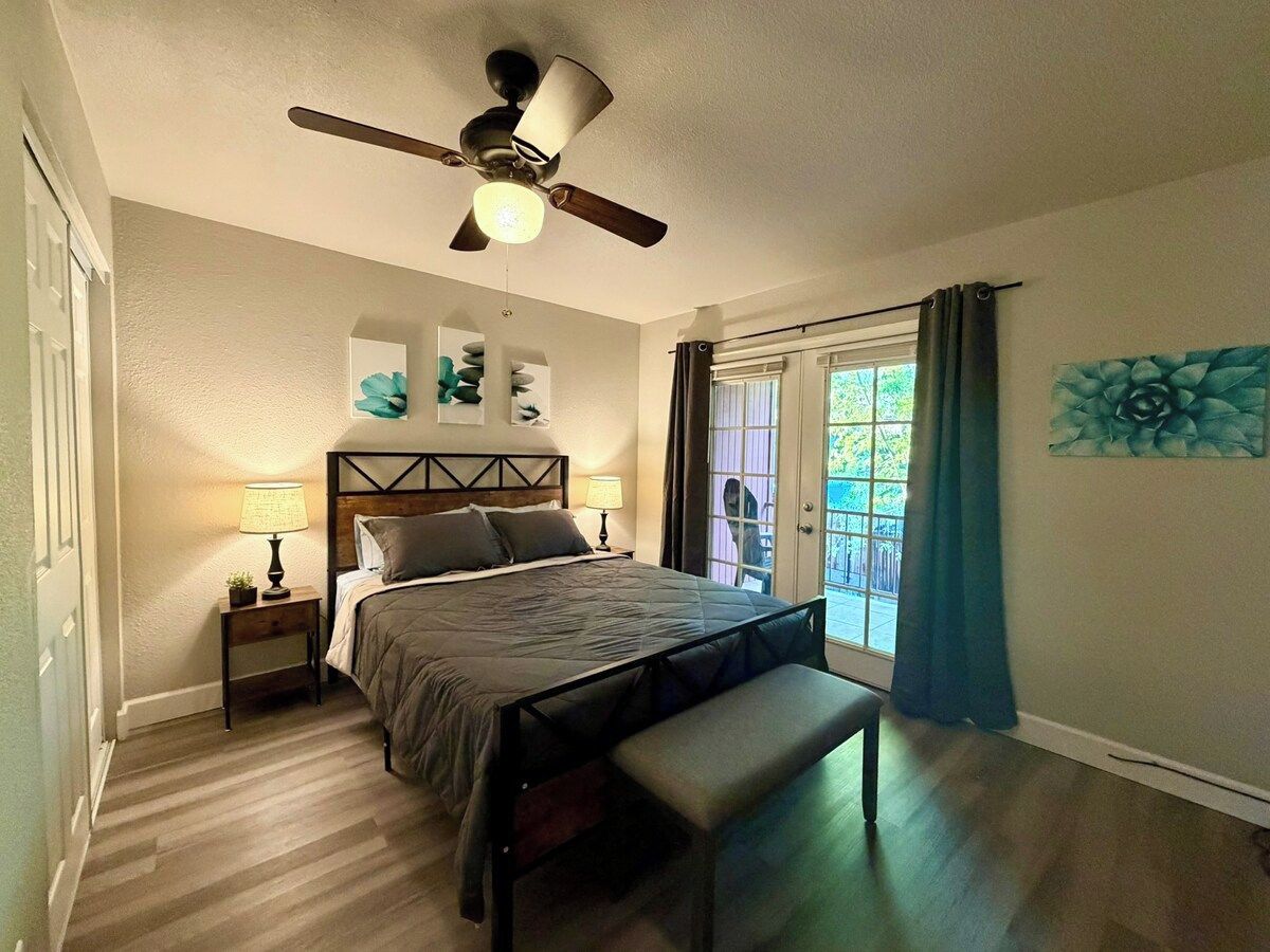 Une chambre à coucher avec un lit, une table de chevet, une lampe, un ventilateur de plafond, un banc et une vue par la fenêtre.