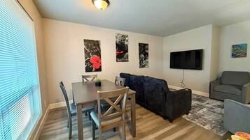 A living room with a dining table, a dark sofa, a grey armchair, and a flat-screen TV mounted on the wall.