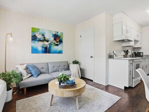 A modern living room with a grey sofa, a round coffee table, and a kitchen area with white cabinets and appliances. - 4 Min Downtown - 7 Min to Duke - 9 Min to Hospital (Durham)