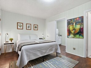 A bedroom with a bed, bedside tables, a nightstand, a painting, and a door leading to another room. - 4 Min Downtown - 7 Min to Duke - 9 Min to Hospital (Durham)