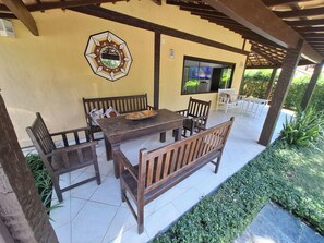 Een overdekt terras met een houten tafel en bankjes, een decoratief wandobject en een televisiescherm.