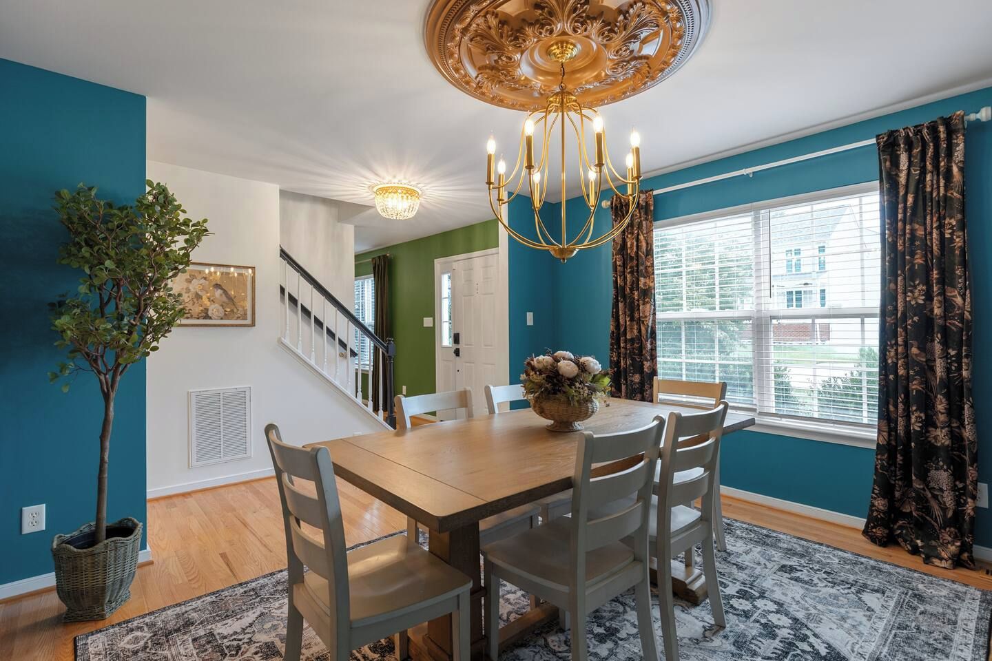 A dining room with a chandelier, a dining table, chairs, a potted plant, and a view of the outside through a window.