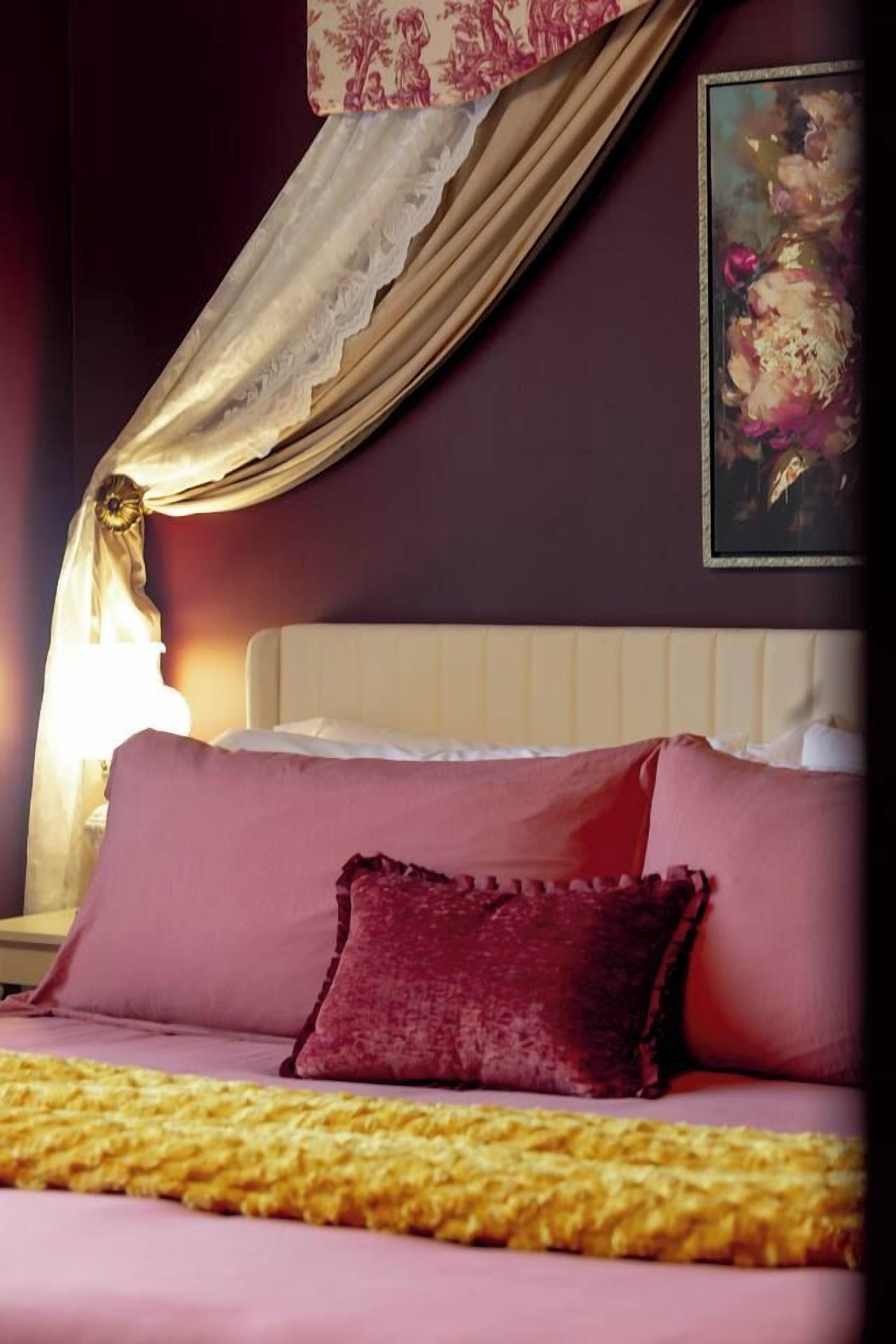 A bed with a pink bedspread and a red pillow, a white headboard, and a painting on the wall.
