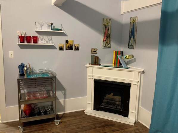 A room with a fireplace, a metal cart with glassware, shelves with decorative items, and framed artwork on the walls.
