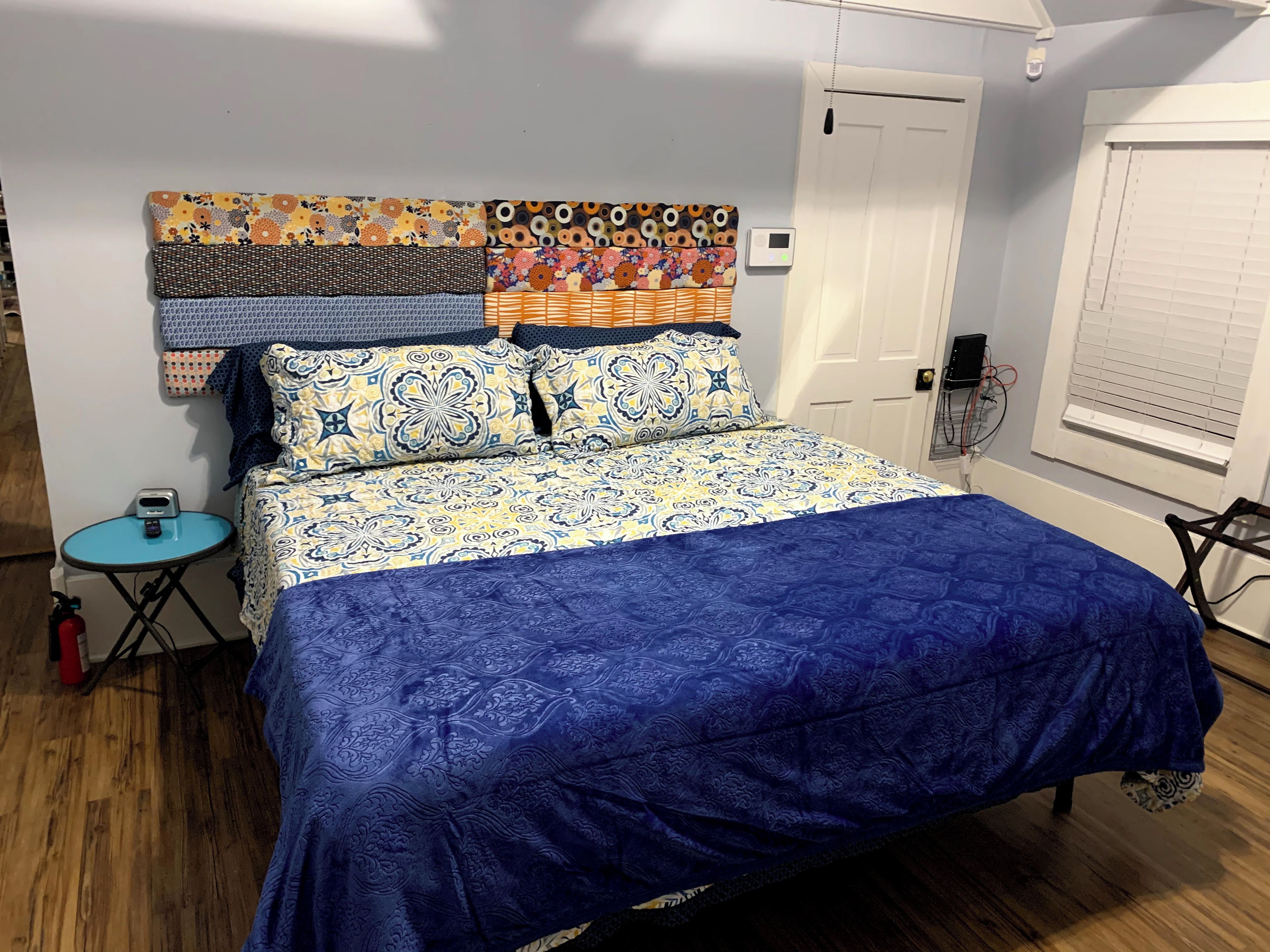 Un lit avec une couvre-lit à motifs et des oreillers, une petite table ronde sur laquelle se trouve un appareil, et un thermostat fixé au mur.
