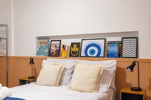 A neatly made bed with two pillows, a bedside table with a lamp, and a shelf above the bed displaying various framed items. - Studio Barão 143  IB03J - Aquarela Stays (Rio de Janeiro)
