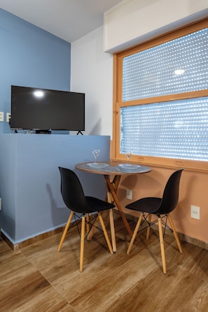 A room with a TV mounted on the wall, a round table with chairs, and a window with blinds. - Studio Barão 143  IB03J - Aquarela Stays (Rio de Janeiro)