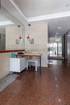 A clean, modern reception area with a reception desk, a chair, and a small table. There are informational signs on the wall, a security camera, and a door leading to another area. - Studio Barão 143  IB03J - Aquarela Stays (Rio de Janeiro)