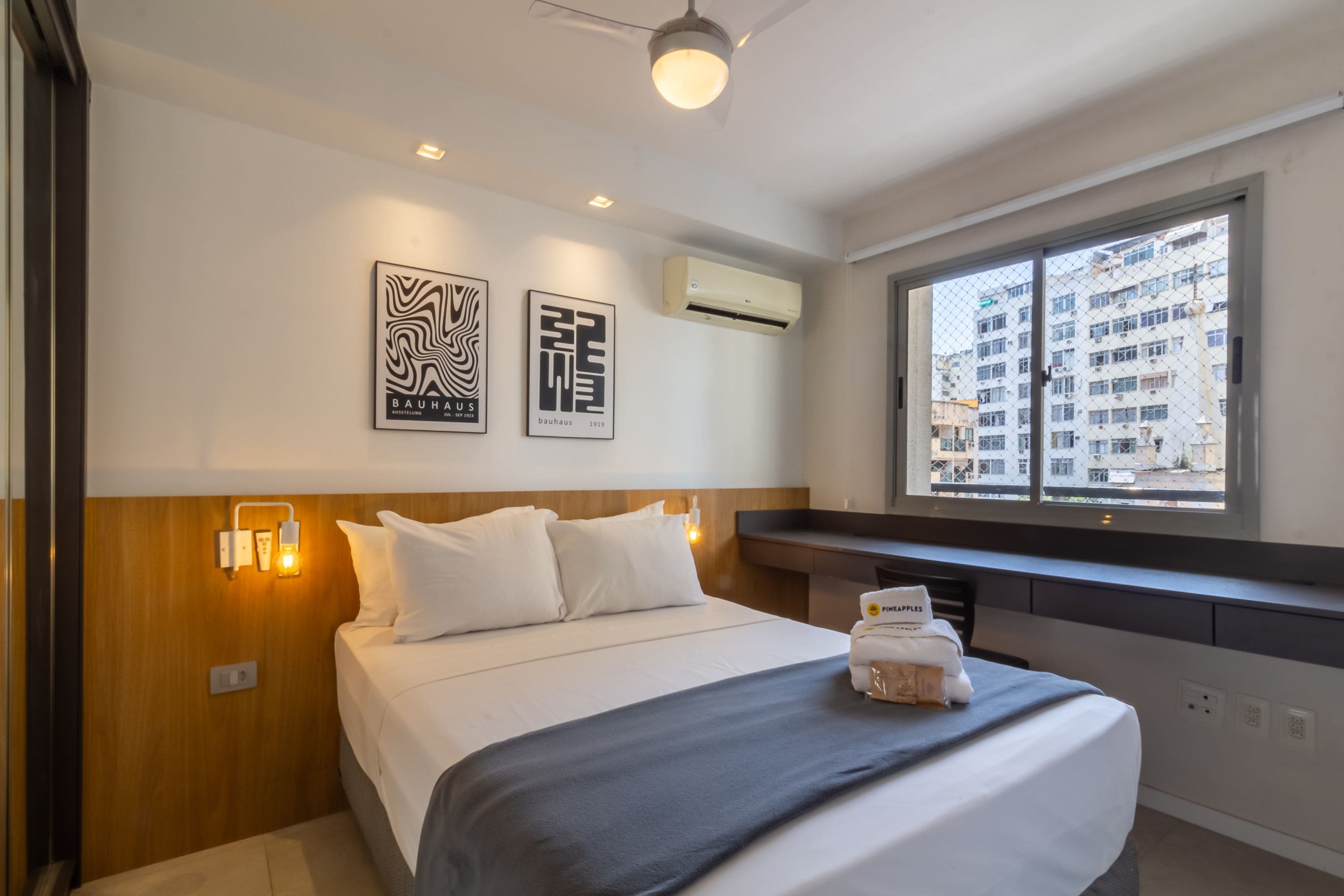 A hotel room with a bed, wall-mounted artwork, an air conditioning unit, and a window with a city view.