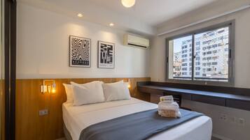 A hotel room with a bed, wall-mounted artwork, an air conditioning unit, and a window with a city view.