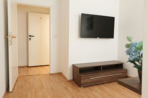 A room with a wooden floor, a TV mounted on the wall, a door with a number, and a plant. - Bring Everyone 3BR near Belvedere Palace (Vienna)