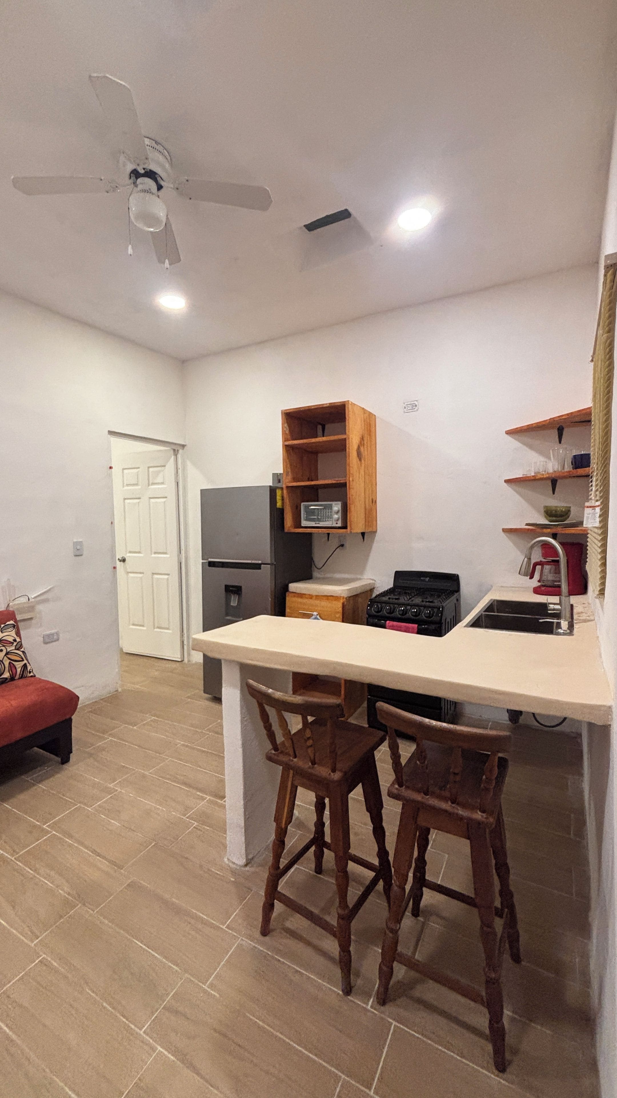 A compact kitchen with a central island, wooden chairs, a refrigerator, a stove, and a ceiling fan.