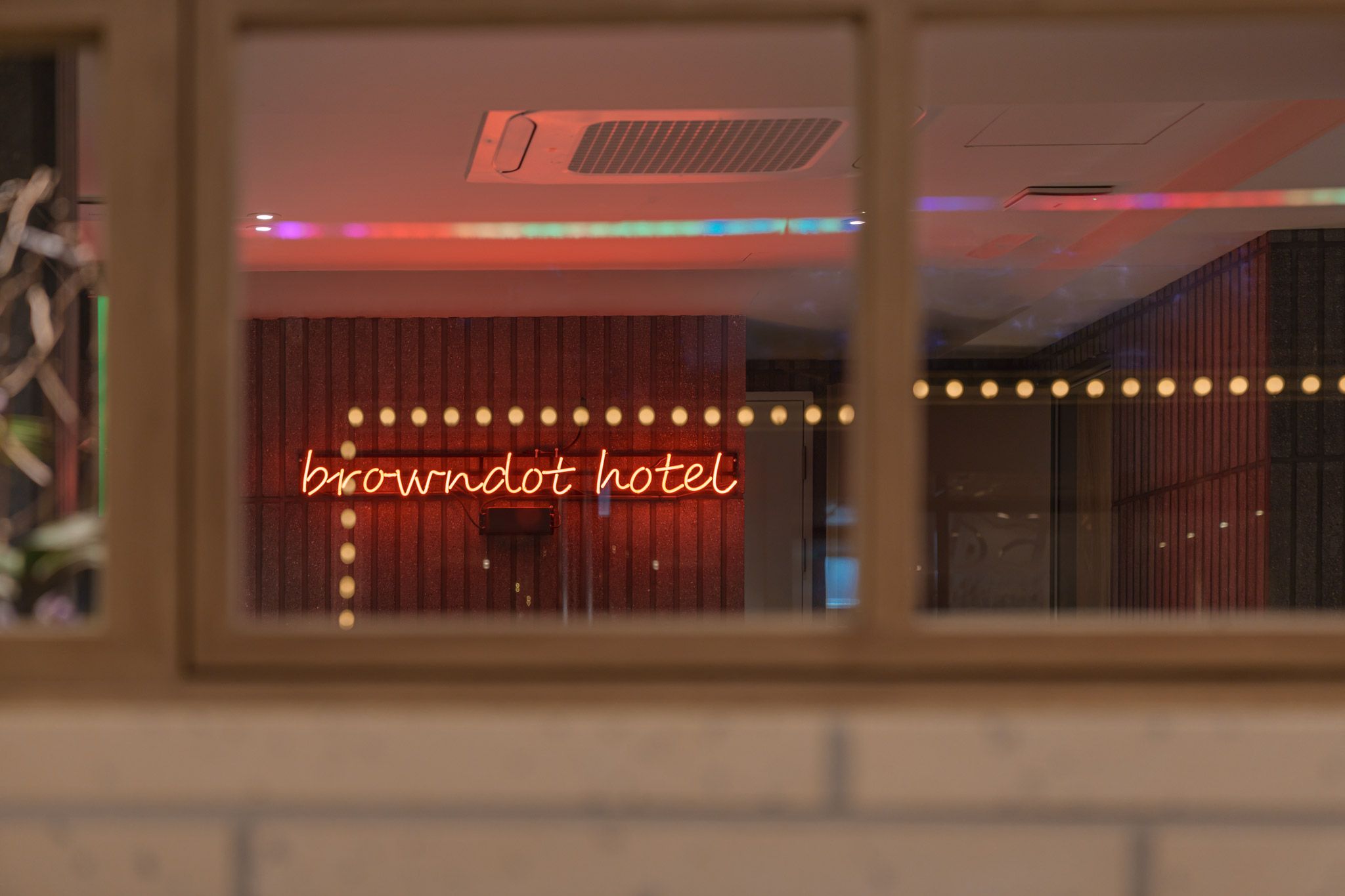 A window view of a room with a neon sign reading 'browndot hotel' and a brick wall.