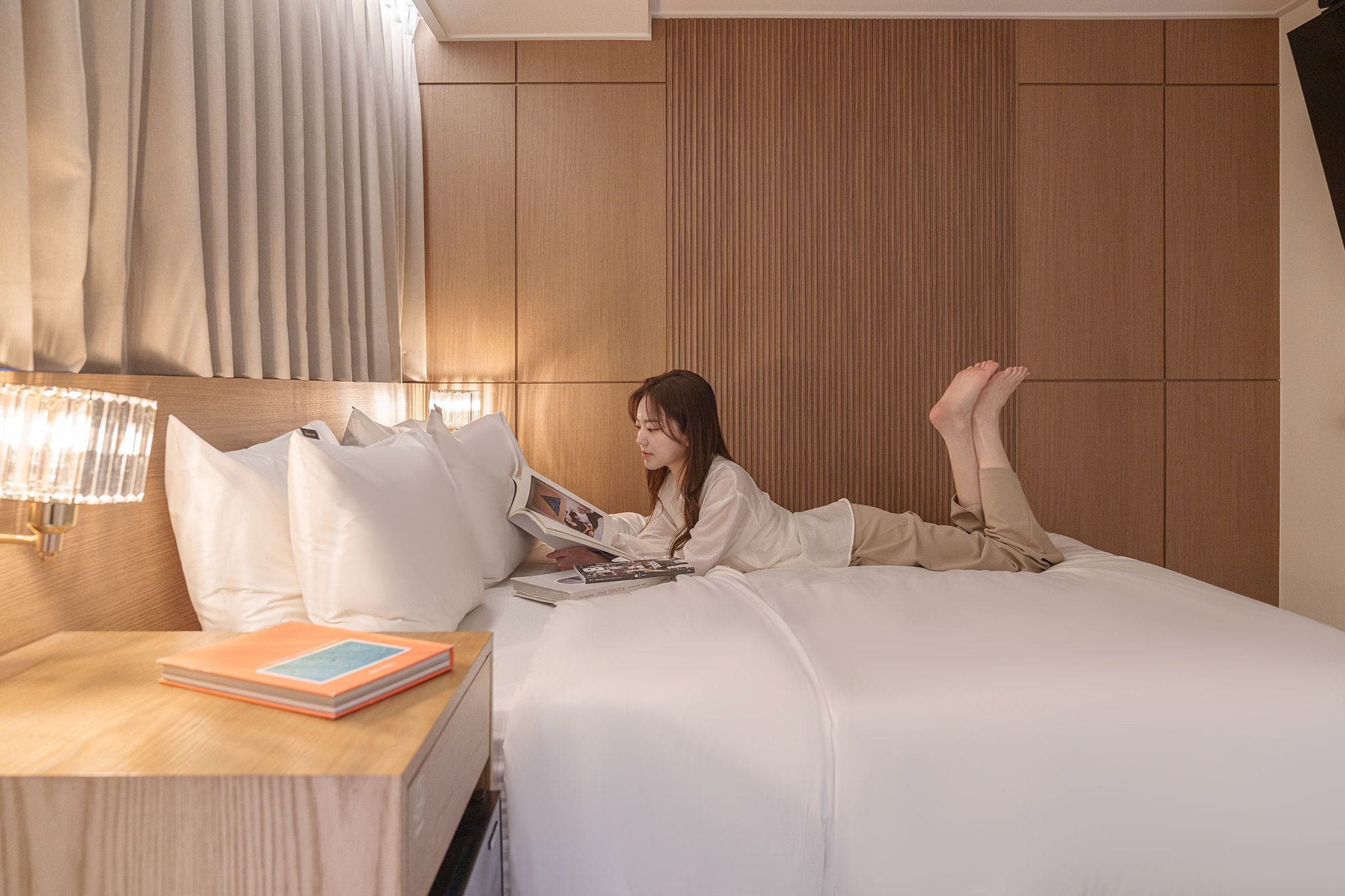 A person lying on a bed in a hotel room, reading a book.
