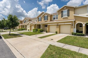 A row of townhouses with attached garages, a well-maintained lawn, and a clear sky. - Next to Disney! 4/4 Townhome, Themed Rooms, 6 Beds (Kissimmee)