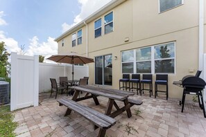 Une terrasse extérieure avec une table et des bancs en bois, un barbecue et une clôture blanche.