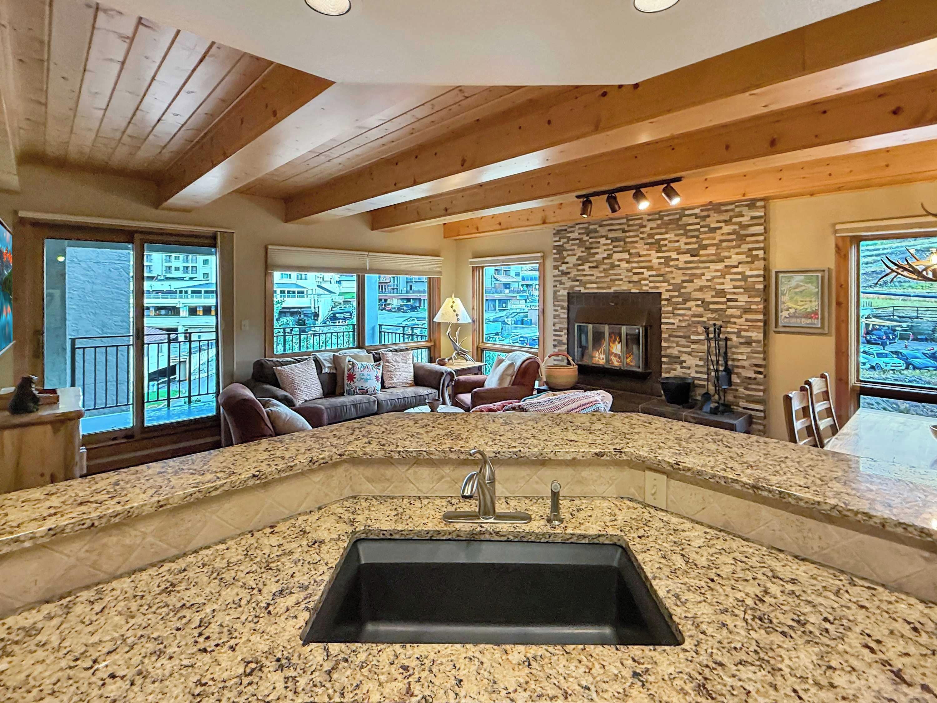 A modern kitchen with a granite countertop and a central sink, a cozy living area with a sofa and a fireplace, and large windows offering a view of the outside.