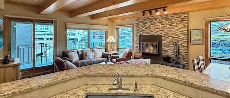 A modern kitchen with a granite countertop and a central sink, a cozy living area with a sofa and a fireplace, and large windows offering a view of the outside.