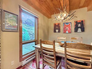 A dining area with a wooden table and chairs, a chandelier, and a window with a view of a mountain landscape. - NEW LISTING - Plaza 2 Bedroom Basecamp—Steps to Trails & Lift! (Crested Butte)