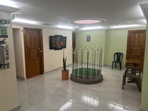 Un hall con suelo de baldosas, puertas de madera, una planta en maceta, una jaula metálica con base verde y un extintor.