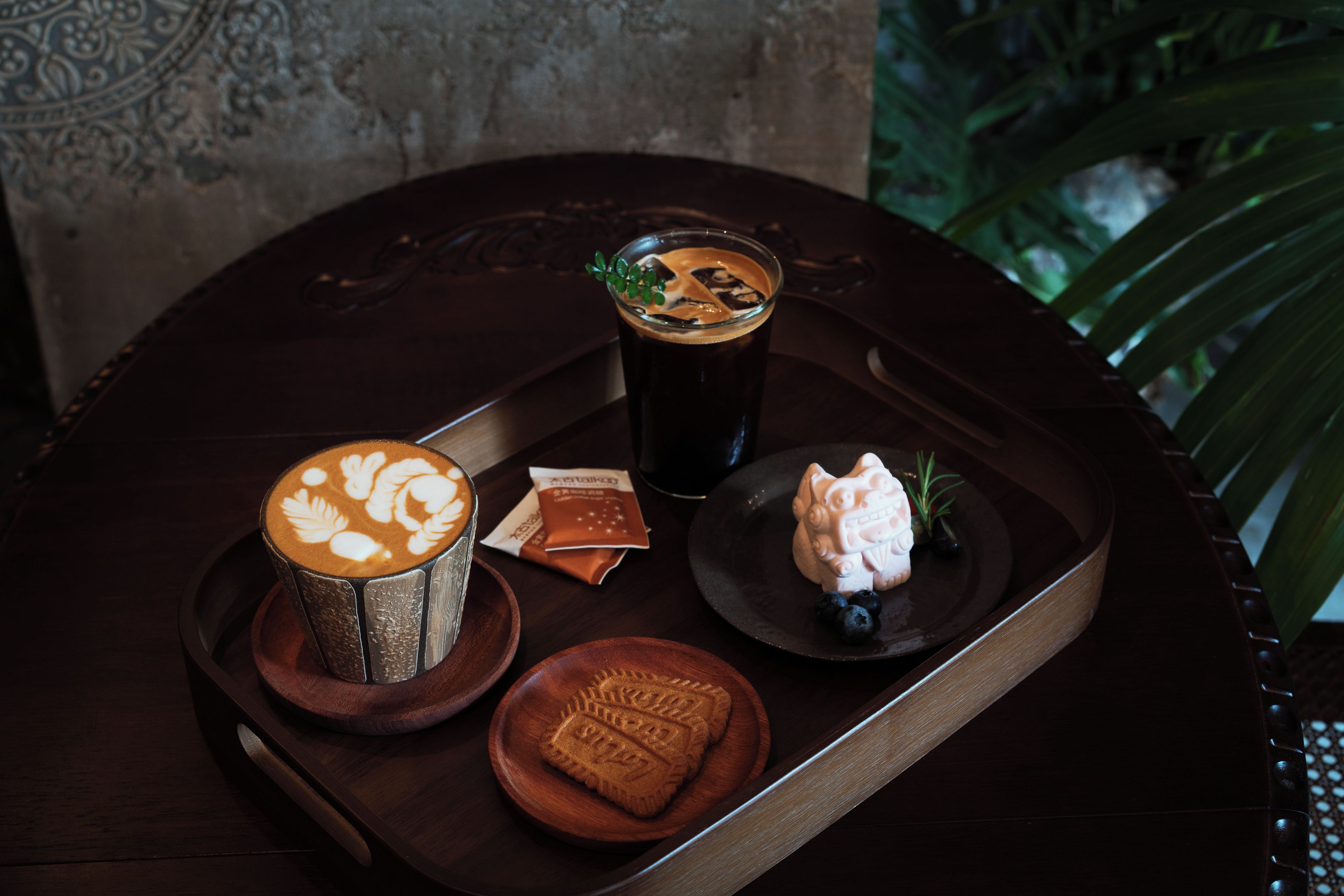 A tray with a coffee cup, a glass of dark beverage, a small figurine, and a cookie on a wooden table.