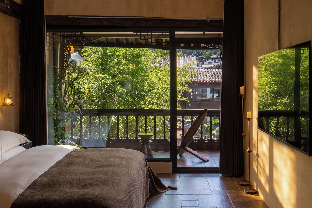 A room with a bed, a wooden chair, a balcony with a view of trees and buildings, and a traditional tiled roof visible through the window.