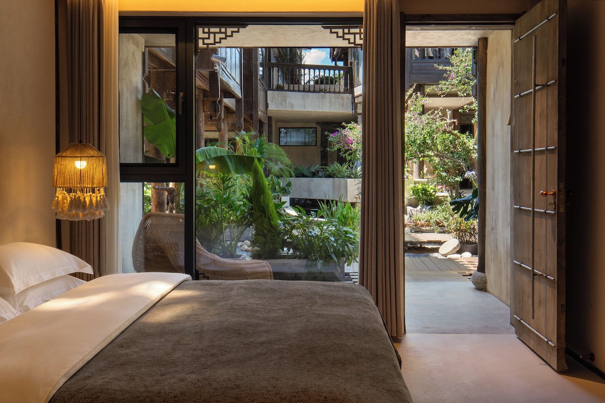 A bedroom with a bed, a hanging light fixture, and a view of a garden through an open door.