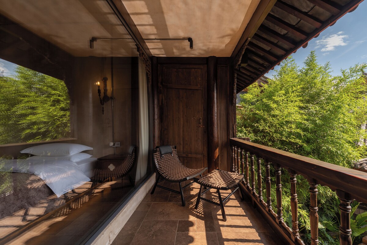 A balcony with a wooden railing, a wicker chair, a cushioned bench, and a view of greenery.