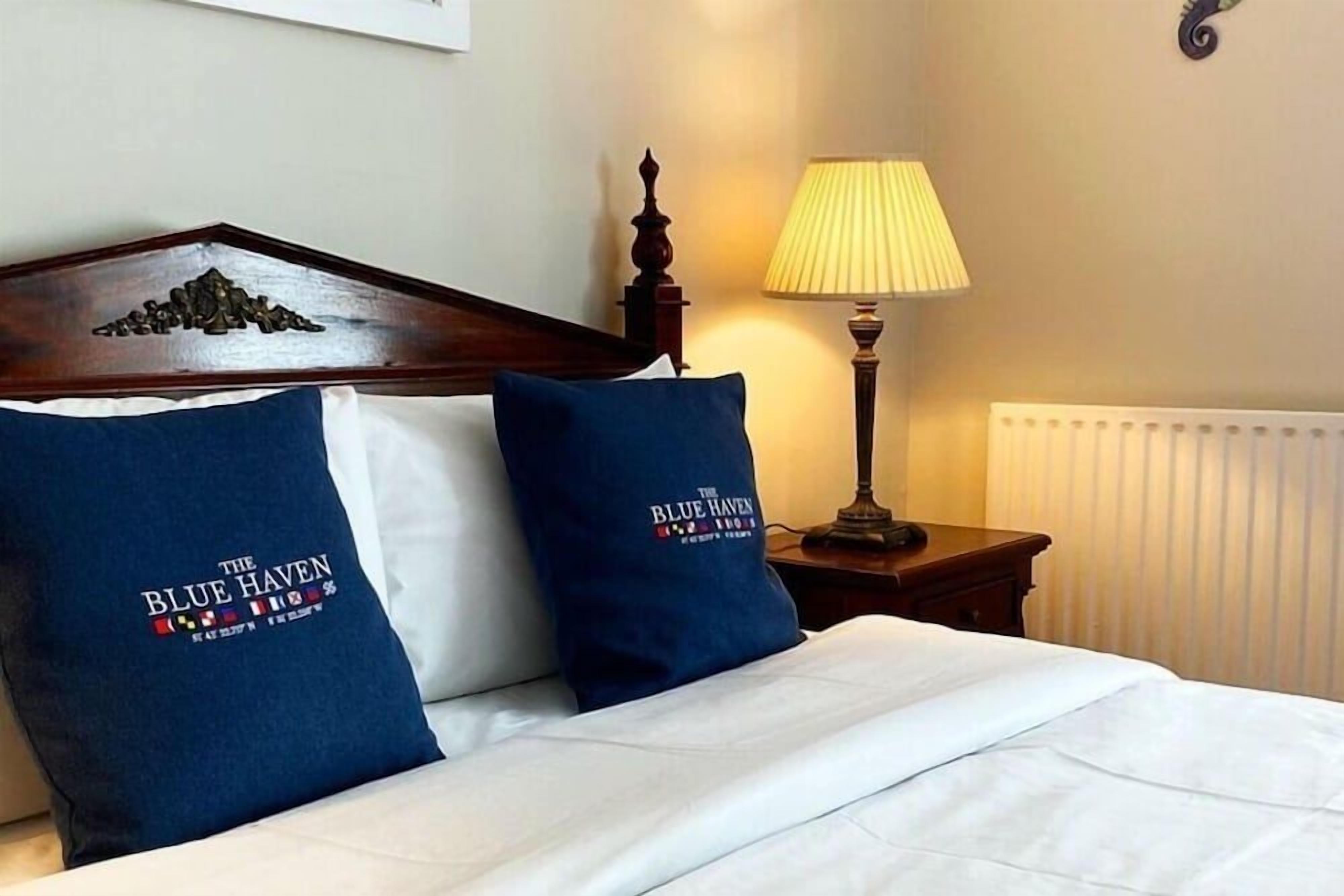 A hotel room with a bed, two pillows with 'The Blue Haven' logo, a wooden headboard, a bedside lamp, and a radiator.