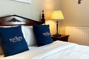 A hotel room with a bed, two pillows with 'The Blue Haven' logo, a wooden headboard, a bedside lamp, and a radiator.