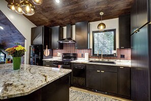 A modern kitchen with dark cabinetry, a granite countertop, and a large window. - Haven Haus Properties (Eagle River)