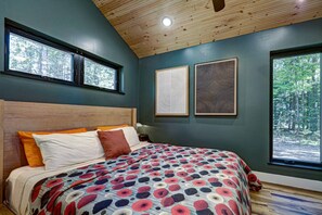 A bedroom with a wooden ceiling, a large bed with a patterned bedspread, two framed artworks, and a view of trees through a window. - Haven Haus Properties (Eagle River)