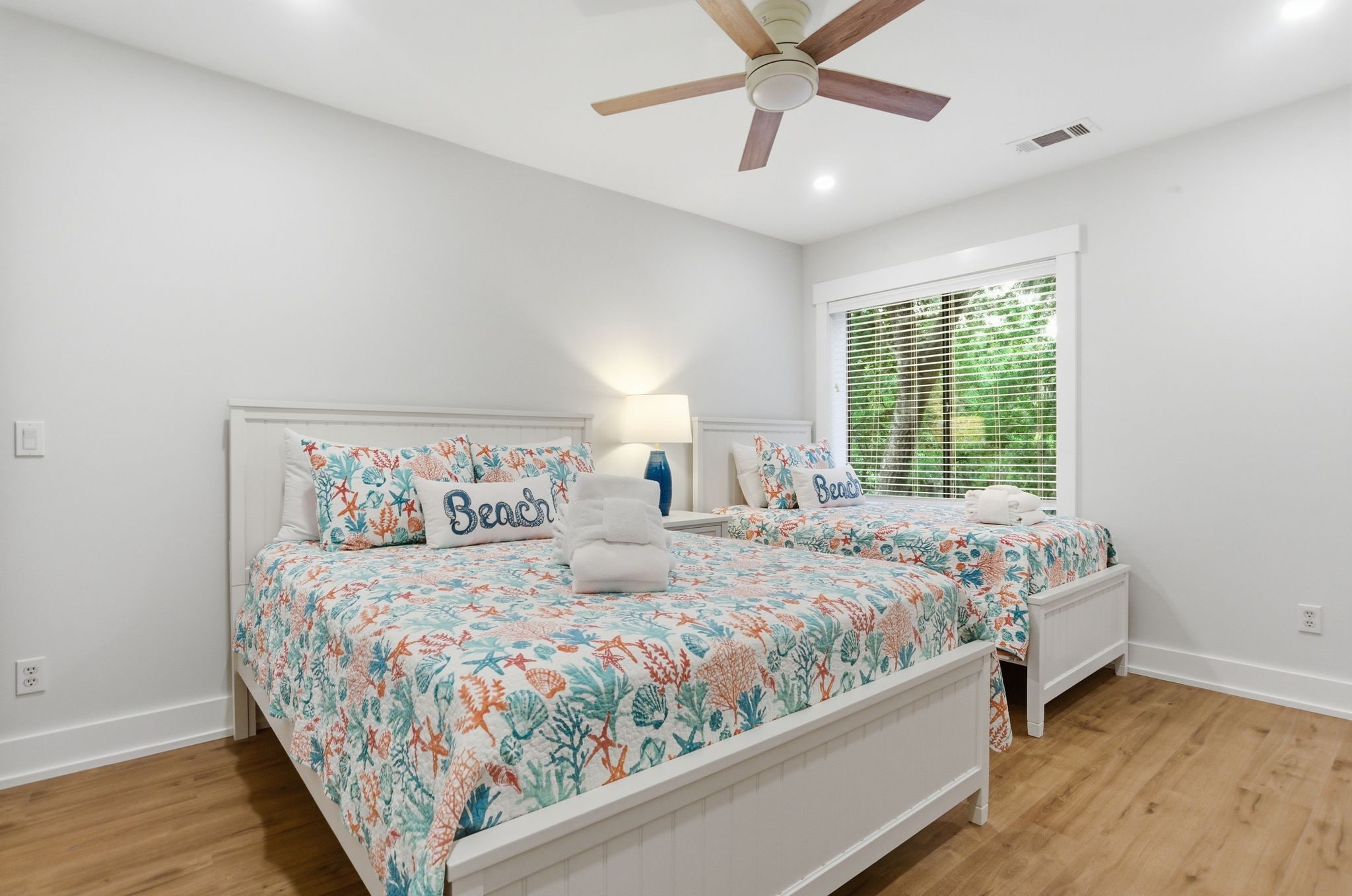 A bedroom with two beds, a ceiling fan, a window with blinds, and a bedside lamp.